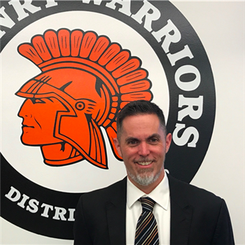 Dr. Jeffry Prickett stands in front of the McHenry Warriors sign, showing his spirit.